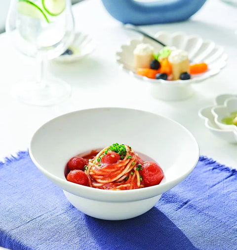 A white ceramic bowl filled with spaghetti in red tomato sauce, topped with cherry tomatoes and green herb garnish, set on a blue napkin on a white table.