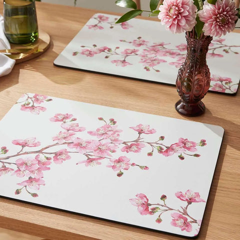Floral placemats on a wooden table with a vase of flowers and a glass.