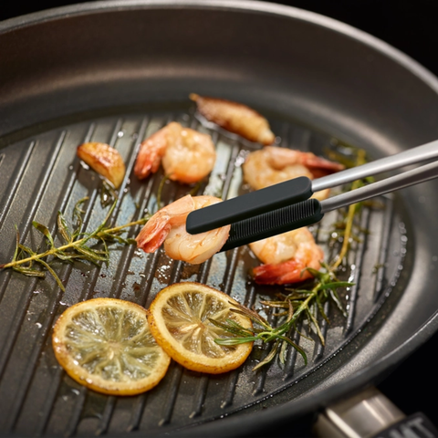 A close-up of a black grill pan with shrimp, lemon slices, rosemary, and oil on a dark background, with metal tongs holding a shrimp over the grates.