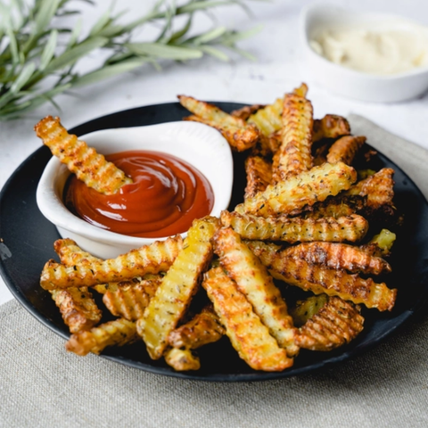 Crispy golden-brown crinkle-cut fries piled on a black plate, served with a small white dish of red ketchup and a small white dish of creamy dipping sauce on a light tabletop.