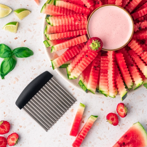 A metal watermelon slice cutter with a black handle is shown on a white surface, with sliced watermelon arranged in a circle and a small bowl of pink drink nearby. Limes, basil leaves, strawberries, and extra watermelon slices are scattered around as styling props.
