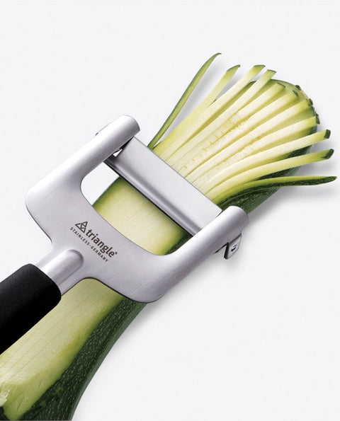 Zucchini being sliced with a silver vegetable peeler on a white background