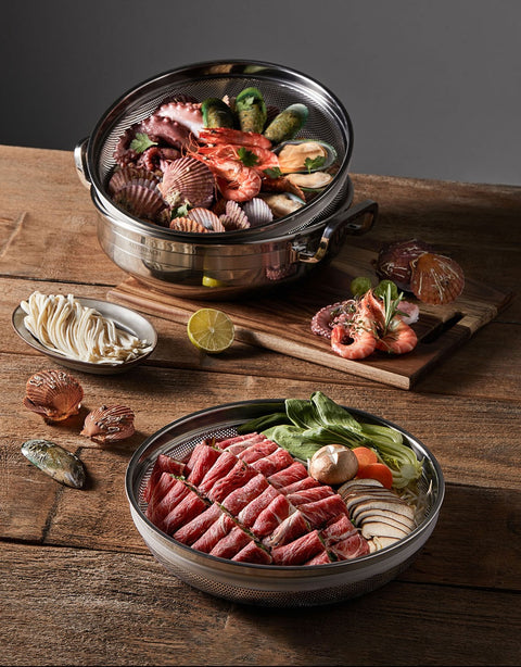 Two metal steamer baskets with raw seafood and vegetables on a wooden surface.
