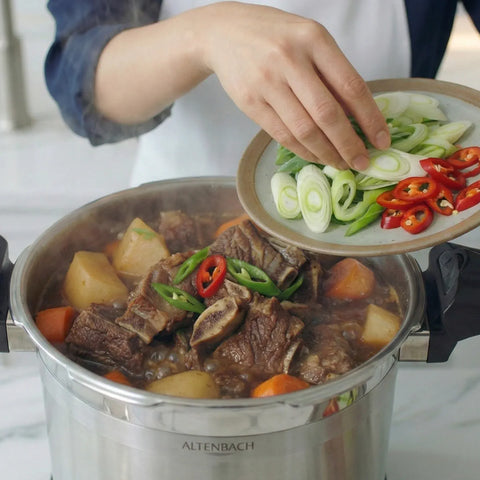 Stewing pot with beef and vegetables, person adding green onions and red peppers.