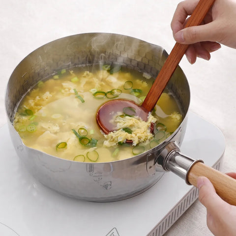 A stainless steel saucepan filled with a light yellow soup, green onions, and egg pieces, being stirred with a wooden-handled spoon on a white electric burner. A hand holds the spoon handle, and another hand holds the pan handle.