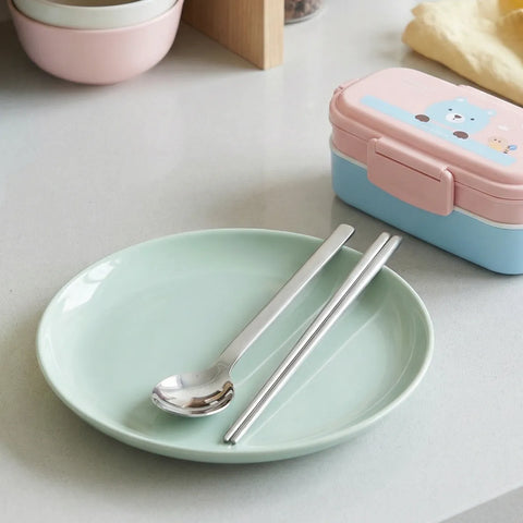 Light green plate with silver spoon and chopsticks, pink and blue bento box on a light surface.