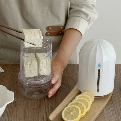 Person holding a container of tofu next to a white appliance on a wooden surface with lemons.