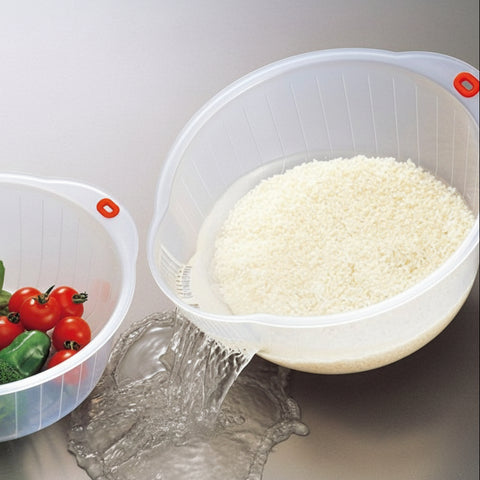 A clear plastic kitchen strainer bowl with a red handle is shown pouring rice into a sink while another bowl with vegetables sits beside it.