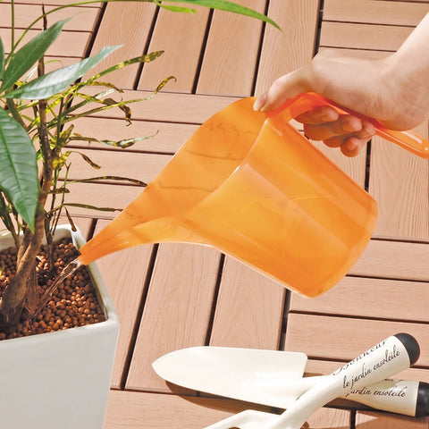 An orange translucent plastic watering can being held by a hand and used to water a potted plant on a brown wooden deck, with white garden trowels visible in the foreground.