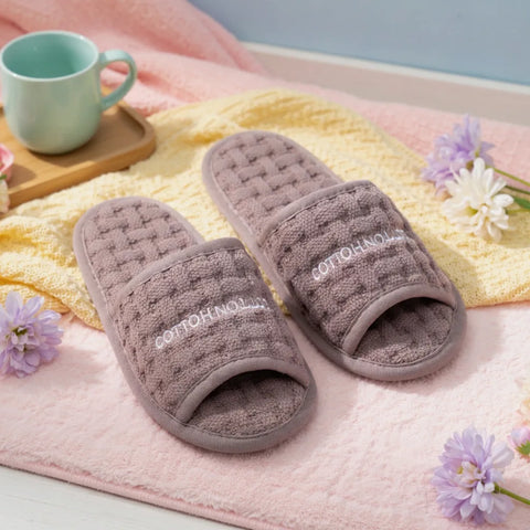 Pair of brown slippers with 'Cotton House' branding on a pink surface with flowers and a mug.
