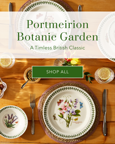 A styled tabletop display of Portmeirion Botanic Garden ceramic plates and bowls with floral and botanical illustrations, arranged on a wooden table with cutlery. A semi-transparent banner across the center reads 'Portmeirion Botanic Garden' with a green 'SHOP ALL' button.