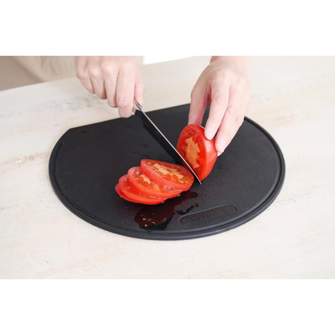 A round black plastic cutting board shown on a light beige countertop. A person's hands are visible slicing a red tomato on the board, which has a small embossed brand logo near the bottom edge.