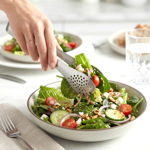 Hands using slotted silver tongs to grab spinach, tomato and cucumber salad on a white table.