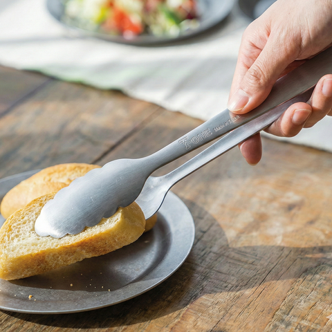 Person grabbing a slice of bread with metal tongs on a wooden table.