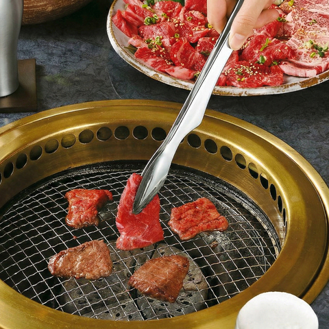 Grilling beef with metal tongs on a small grill with a plate of sliced beef in the background.