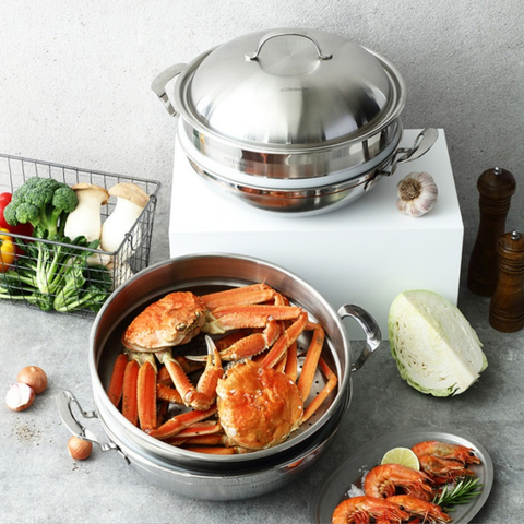 A stainless steel multi-tier steaming pot set displayed on a gray stone surface with food props. The top pot with lid sits on a white block, while the lower pot contains cooked crab meat and carrots.