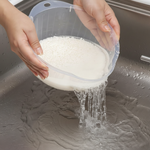 A clear plastic washbowl being held by two hands over a stainless steel sink, with white rice inside and water draining into the sink.