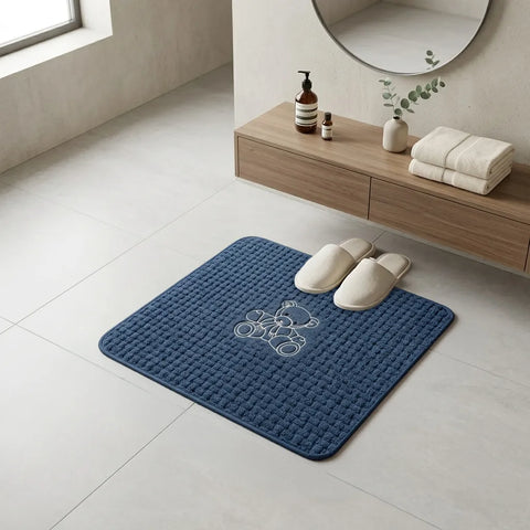 Blue bath mat with a logo on a bathroom floor, next to white slippers and a wooden shelf with towels and bottles.
