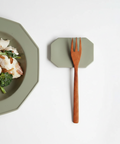 A wooden pasta fork with a brown handle rests on a muted sage green octagonal magnetic stand, next to a matching sage green octagonal plate containing food, all on a clean white background.
