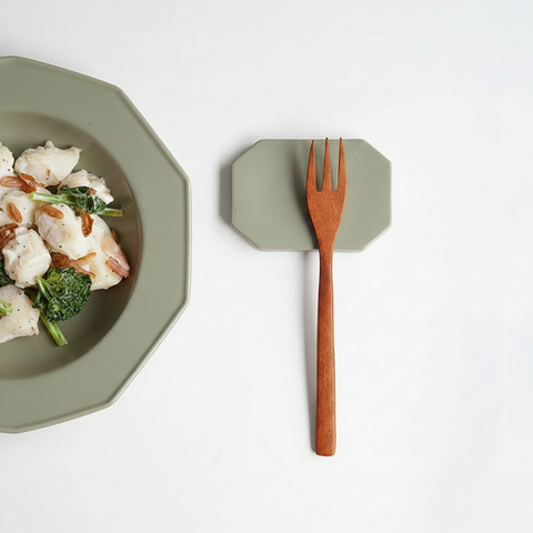 A wooden pasta fork with a brown handle rests on a muted sage green octagonal magnetic stand, next to a matching sage green octagonal plate containing food, all on a clean white background.