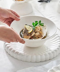 A white ceramic soup bowl with a fluted rim, being held by two hands over a matching scalloped white plate. The bowl contains a piece of fish in broth with a lemon wedge and parsley garnish, set on a white tablecloth.
