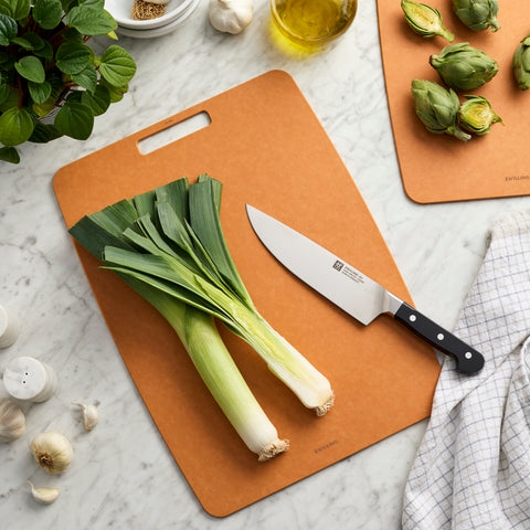 Brown cutting board with leeks and a knife on a marble countertop.