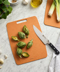 Brown cutting board with green vegetables and a knife on a marble surface.