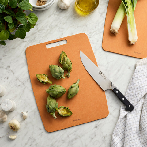 Brown cutting board with green vegetables and a knife on a marble surface.