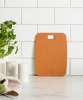 Brown cutting board on a white surface with green herbs and a plant in the background.