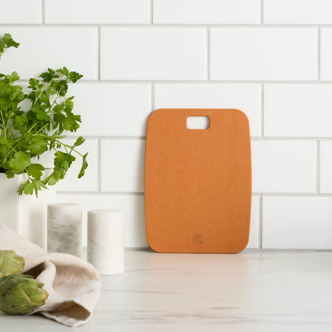 Brown cutting board on a white surface with green herbs and a plant in the background.