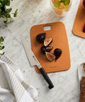 Brown cutting board with figs and a knife on a marble surface, surrounded by bread and a plant.