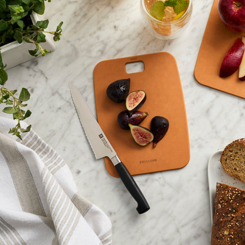 Brown cutting board with figs and a knife on a marble surface, surrounded by bread and a plant.