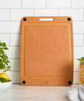 Brown cutting board on a kitchen counter with a glass of lemonade and a bowl of fruit.