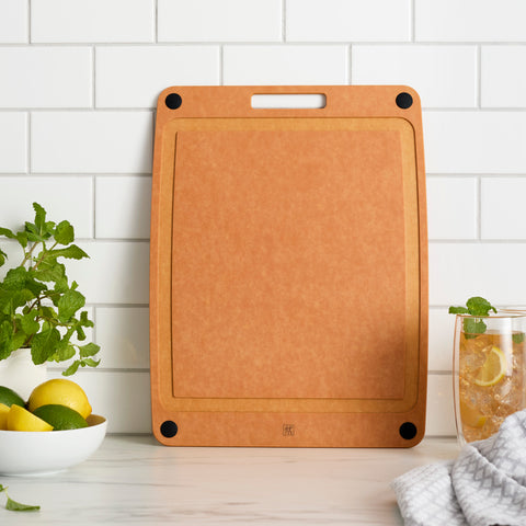 Brown cutting board on a kitchen counter with a glass of lemonade and a bowl of fruit.