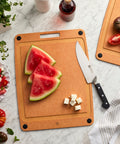 Wooden cutting board with watermelon slices, knife, and vegetables on a marble surface
