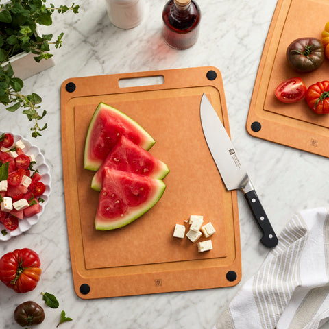 Wooden cutting board with watermelon slices, knife, and vegetables on a marble surface