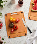 Wooden cutting board with tomatoes and a knife on a marble surface, next to a plate of salad and watermelon.