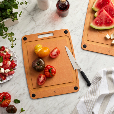 Wooden cutting board with tomatoes and a knife on a marble surface, next to a plate of salad and watermelon.