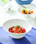 A white ceramic bowl filled with spaghetti in red tomato sauce, topped with cherry tomatoes and green herb garnish, set on a blue napkin on a white table.