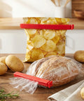 Red plastic bag clip on a bag of potato chips and bread on a wooden table with potatoes around.