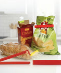Red bag clips used on a bag of chips and a box of crackers on a kitchen counter.