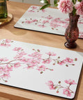 Floral placemats on a wooden table with a vase of flowers and a glass.