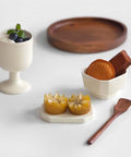 A styled tabletop arrangement of ceramic tableware on a white surface: a small off-white pedestal bowl with blueberries and mint, a white dish holding a round pastry and a brown chocolate piece, two small yellow fruit pieces on a white tray, a wooden spoon, and a round wooden plate in the background.