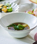 A white ceramic deep bowl containing a clear broth with green seaweed and leafy greens, set on a white tablecloth with sushi and a salad blurred in the background.