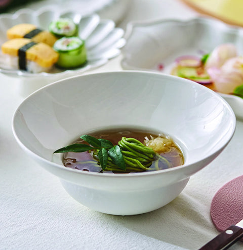 A white ceramic deep bowl containing a clear broth with green seaweed and leafy greens, set on a white tablecloth with sushi and a salad blurred in the background.