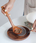 A person's hands are shown using a wooden spoon to strain a brown paste over a small metal sieve set on a round wooden plate, with a white plate in the background.