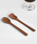 Two wooden spoons with a warm medium-brown finish, arranged diagonally on a clean white surface with a glass base partially visible in the upper right.