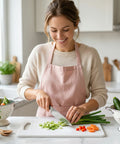 Woman in a kitchen wearing a pink apron, chopping vegetables on a cutting board.