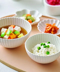 White ribbed ceramic bowls arranged on a peach-colored tray, containing a vegetable tofu soup and white rice with green peas, with small side dishes in the background.