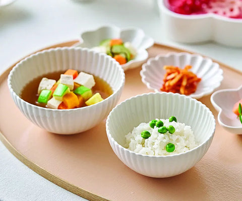 White ribbed ceramic bowls arranged on a peach-colored tray, containing a vegetable tofu soup and white rice with green peas, with small side dishes in the background.